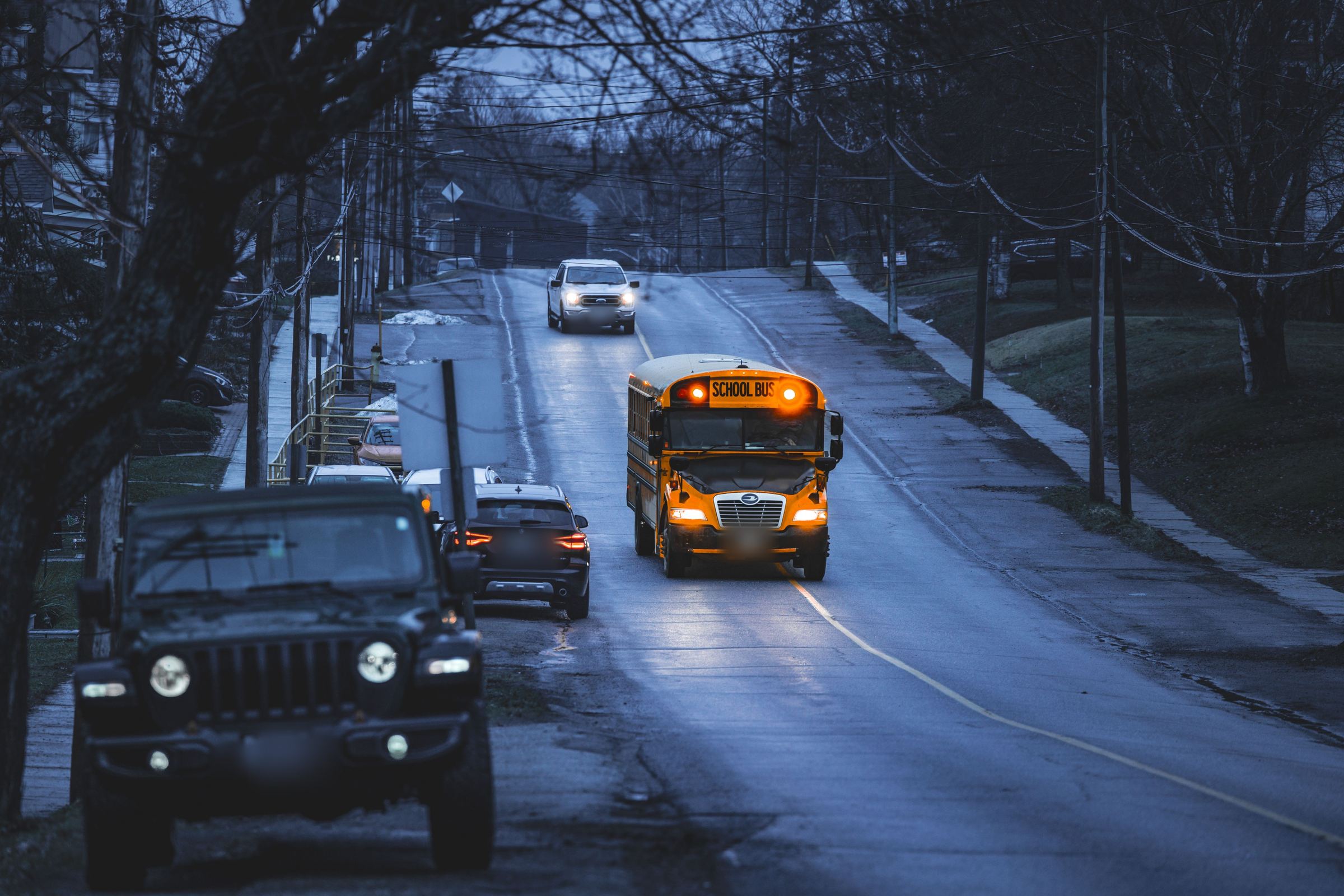 A school street at dusk, a yellow school bus with its lights on